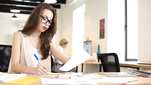 Young business woman making notes in notebook while sitting at the table in office with her colleague on the background