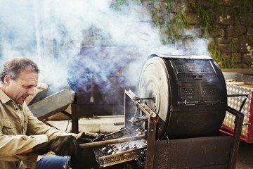 A man roasting chestnuts over hot coals in a large drum, raking out the roasted nuts. 