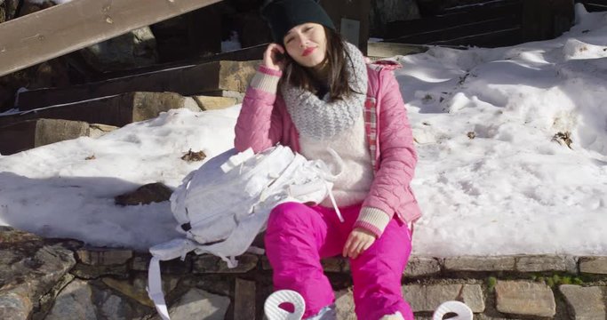 Young beautiful asian woman relaxing on whe wooden bench in snowy winter conditions. She wearing pink ski clothes. Smiling to the camera.