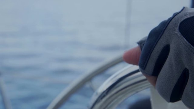 A sailor is holding a steering wheel on a sailing boat. He is wearing protection gloves. We can see a peaceful sea and a few islands in the background.
