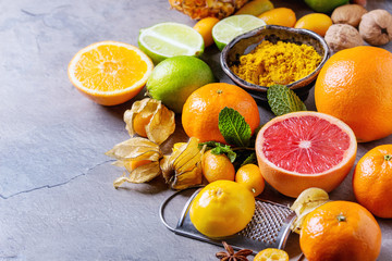 Variety of whole and sliced citrus fruits pineapple, grapefruit, lemon, lime, kumquat, clementine and physalis with mint and yellow spices and nuts over gray textured background. Copy space. 