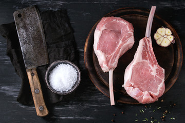 Two raw uncooked Veal tomahawk steak with garlic, salt, seasoning on wooden chopping board and vintage butcher cleaver on textile napkin over black texture background. Top view with space.