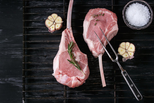 Two raw uncooked Veal tomahawk steak with garlic, salt and seasoning on old grill grate with meat fork over black texture background. Top view with space.