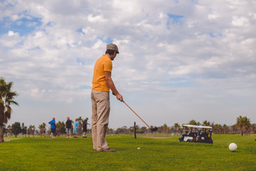 Back view of active man holding playing vintage wooden golf club on palm tree grass field outdoors background.