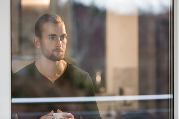 Lonely man wearing a black long sleeved shirt, standing in his home looking out of the window holding a coffee cup. 