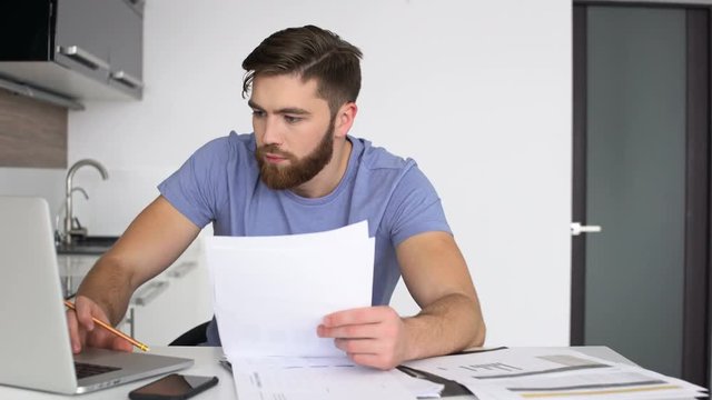 Young bearded man working with laptop and documents while his wife bringing him cup of coffee and hugging 