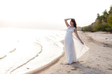 Beautiful young brunette woman in white long dress at the sea shore