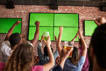 Rear View Of Friends Watching Game In Sports Bar On Screens