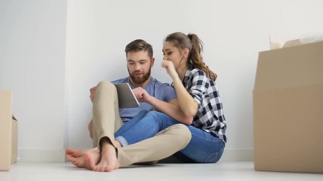 Young happy couple with digital tablet sitting on the floor in their new home