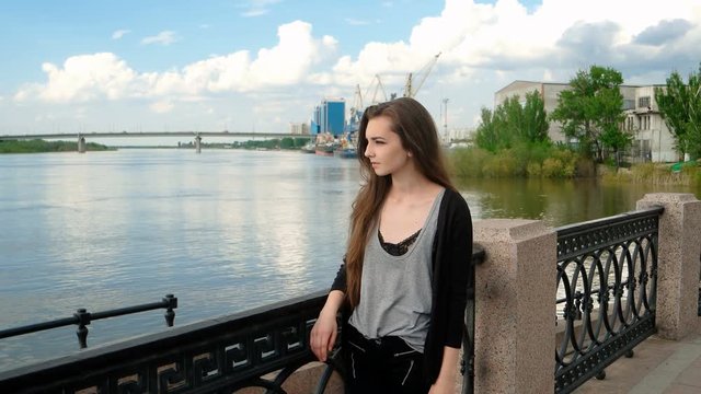 Young beautiful lady waiting on the embankment wearing gray cashmere jacket. Young girl standing ner fence in waterfront area in pencive state..