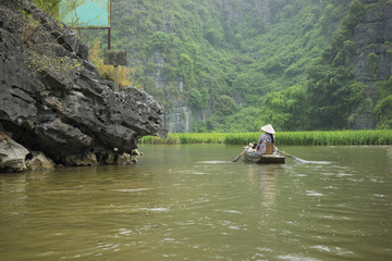 Vietnamese woman in traditional conical hat rows boat into natural cave on Ngo Dong river, Tam Coc, Ninh Binh, Vietnam