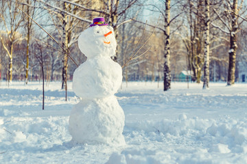 Snowman with purple hat in city park. Evening time