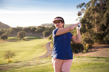 Mature woman playing golf. Golfer hitting golf shot with driver club on course. Beautiful sunny Landscape, green hills, blue sky. Portugal. 