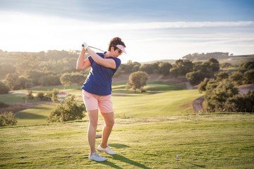 Mature woman playing golf. Golfer hitting golf shot with driver club on course. Beautiful sunny Landscape, green hills, blue sky. Portugal. 