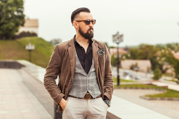 Young confident hipster man with beard in glasses posing on the street in old town. Cute man wearing a vest, black shirt and light trousers. Man keeps hands in pockets and looking forward