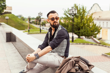 Obraz premium Young confident hipster man with beard in glasses posing on the street in old town. Cute man wearing a vest, black shirt and light trousers. Man keeps hands in pockets and looking forward