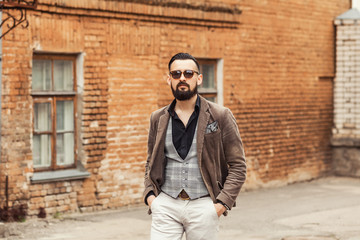 Young confident hipster man with beard in glasses posing on the street in old town. Cute man wearing a vest, black shirt and light trousers. Man keeps hands in pockets and looking forward