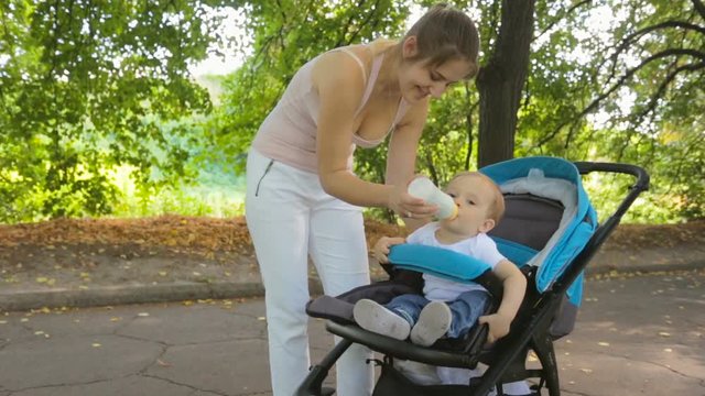 Beautiful woman feeding her baby son from bottle in pram at park