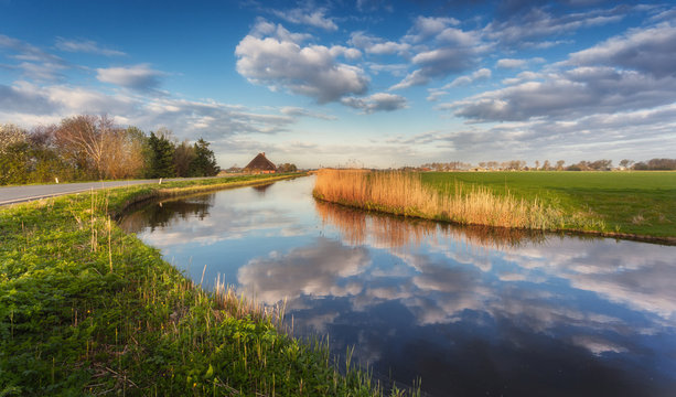 Buildings and trees near the water canal at sunrise in Netherlands. Colorful blue sky with clouds. Spring landscape in Holland. Rural scene. Cloudy sky reflected in water. Nature background - Powered by Adobe