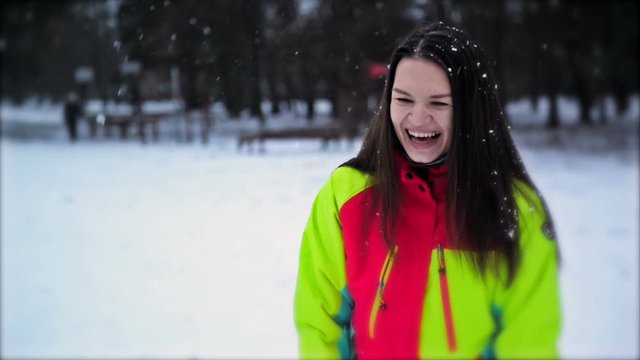 Cute Young Girl with Long Hair and Wonderful Smile Enjoying Winter Outdoors. Closeup Portrait of Woman Wearing Colorful Ski Suit, Snowflakes Slowly Falling Down.