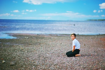 A young girl with short hair in a white blouse and black pants is sitting on his knees on the beach on the background of beautiful blue sky.