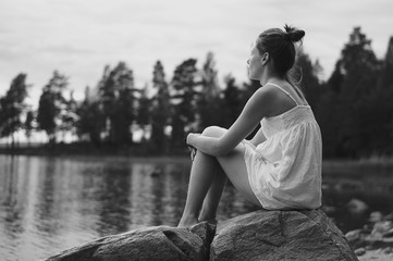 Young woman in white dress sitting on stone at lake contemplating