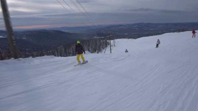 Woman snowboarder is riding dowm fast from snow-covered mountains slope at cloudy day