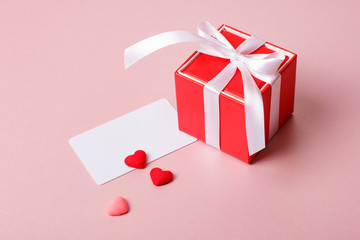 Valentine day composition: red gift box with bow, credit / visiting card template and small hearts on light pink background.