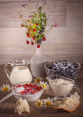On the table strawberries and blueberries in a bowl. In a glass container with milk and sour cream. The pieces of bread on the table. In a vase bouquet of berries.