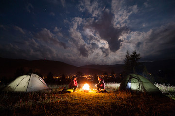 Fototapeta na wymiar Girl and guy sitting at a bonfire near tents in the evening under incredibly beautiful a cloudy sky in the background mountains and luminous town