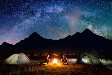Man and woman sitting at a bonfire near tents in the evening under incredibly beautiful starry sky in the background high mountains and luminous town