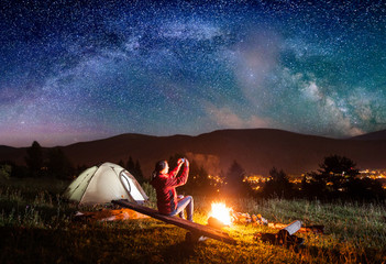 Hiker woman sitting on boards by a campfire near tent and makes the photo incredible view of night sky with stars and Milky way on the phone