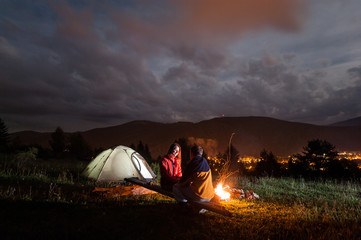 Romantic night for couple of sitting face to face on boards near campfire and tent under cloudy sky on the background mountains and luminous town in the evening