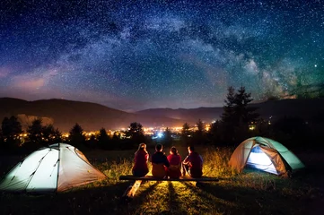 Fotobehang Kamperen Vrienden wandelaars zitten op een bankje gemaakt van boomstammen en kijken samen naar vuur naast kamp en tenten in de nacht. Op de achtergrond prachtige sterrenhemel, bergen en lichtgevende stad. Achteraanzicht