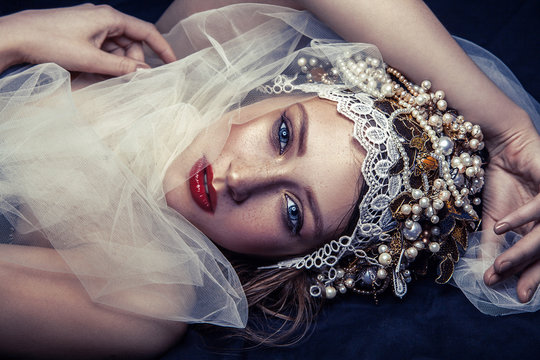 fashion beauty portrait of young beautiful young woman with makeup and freckles on her face and pearl headpiece on her head and white tulle in front of her face on dark blue background. 
