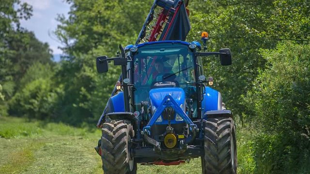 A young farmer is coming on a big lawn with his blue tractor. A grass cutting machinery is connected to it.
