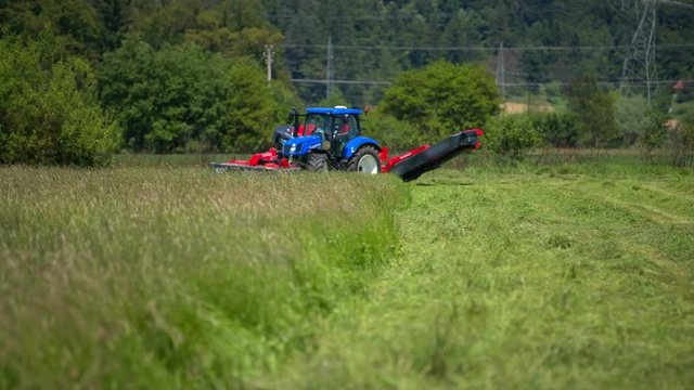 We can see a big blue tractor and a huge grass cutting machinery connected to it. A farmer is starting to cut the grass.
