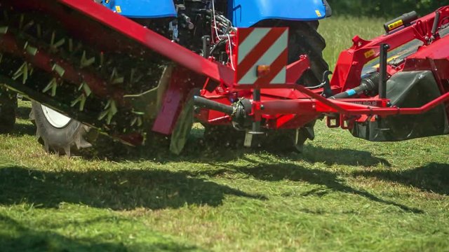 A blue tractor and a red machinery are driving across the lawn and the machinery suddenly lifts up.
