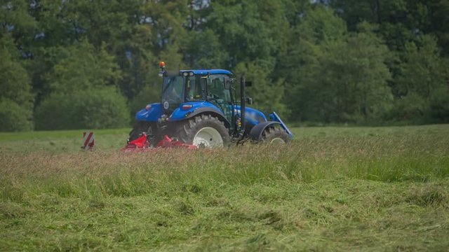 A blue tractor is driving very fast across the grass field. He is going to start working and turning freshly cut grass around.
