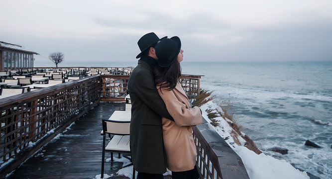 Man embracing his wife standing on the terrace by the sea.Outdoor,fall portrait,fashionable couple,trendy outfit.Two lovers,sea background.hipster.couple in jeans,warm coat,sneakers.Winter holidays