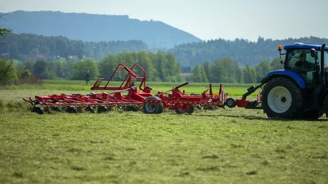 A blue tractor is working and preparing hay on a hot summer day. The big agricultural machinery is turning hay around.
