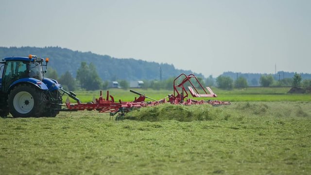 A tractor is lowering down rotary hay rakes and the machinery starts turning hay around which flies around. It is a hot summer day.
