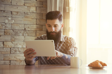 bearded man drinking morning coffee or tea with digital tablet