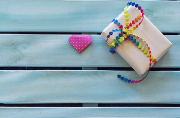 Overhead of handmade gift with vibrant bow tie and origami heart on blue wooden background and copy space. Subjects captured against soft window lighting
