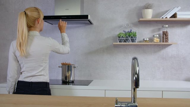 A young woman is standing in front of a kitchen hood in the kitchen and she is carefully pressing a few buttons on the device.
