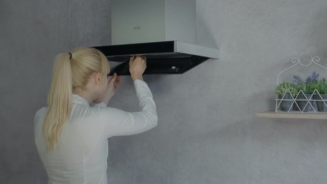 A young woman is opening the lower part of the kitchen hood and then she is taking out a filter.
