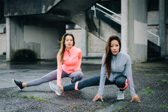 Two sporty women stretching long adductor for warming up before urban fitness workout or running training. Latin and caucasian female athletes doing leg stretch exercise outside on rainy winter day.