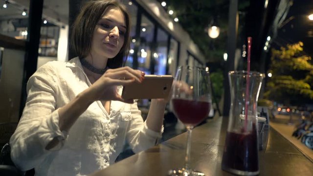 Young woman taking photo of decanter with red wine sitting in cafe at night 
