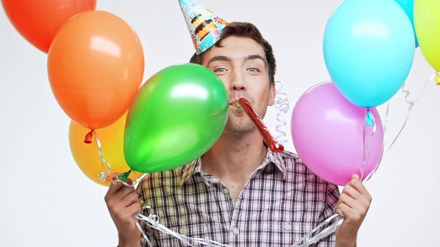 Funny Young Caucasian male with dark hair and light bristle wearing birthday cap cheerfully hiding behind multicolored balloons on white background in slowmotion
