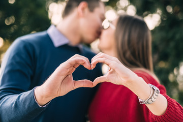 Young, happy couple enjoying outdoors. They making heart shape with hands and kissing. Selective focus on hands.